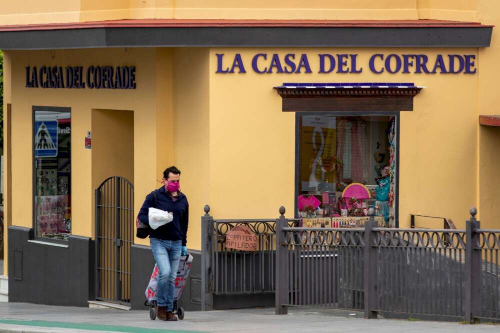 Un hombre protegido con mascarilla camina con un carrito de la compra ante un comercio cerrado de artículos cofraderos en el sevillano barrio de Triana. Foto: Efe