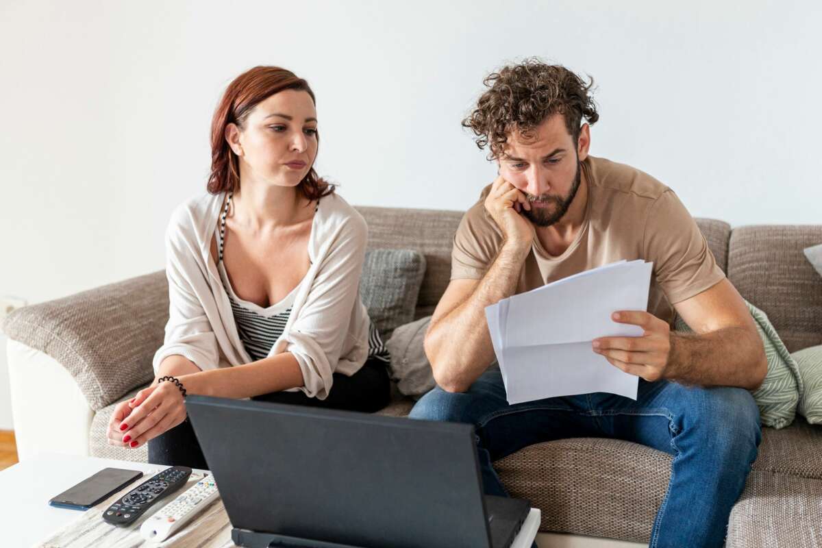 Una pareja consulta preocupada unos papeles sentada en el sofá de su casa mientras realiza una gestión con el ordenador. Foto: Freepik.