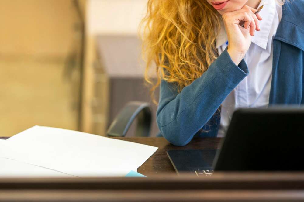 Una mujer consulta un papel mientras realiza un trámite con un ordenador. Foto: Freepik.