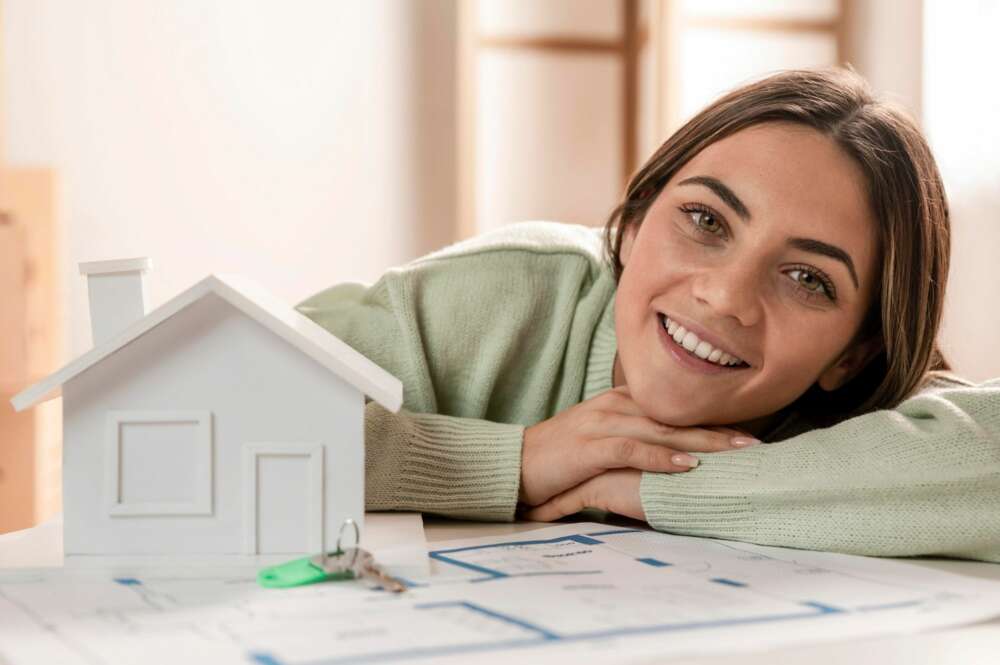 Una joven recostada en sus manos con una maqueta y los planos de una casa. Foto: Freepik.