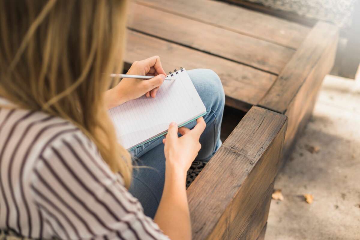 Una persona escribe en su libreta sentada en un banco de madera. Foto: Freepik.