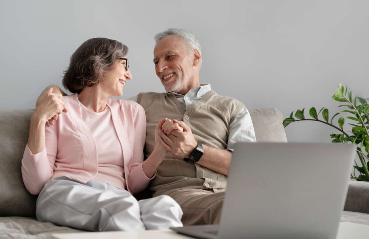 Una pareja de personas mayores se dan la mano mientras realizan una consulta con el ordenador. Foto: Freepik.