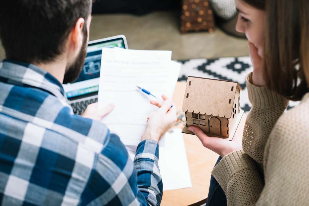 Una pareja consultan unos papeles mientras sostienen una casa de manera. Foto: Freepik.