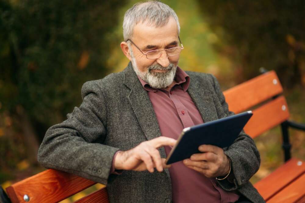 Hombre mayor leyendo en su tablet en la banca de un parque.