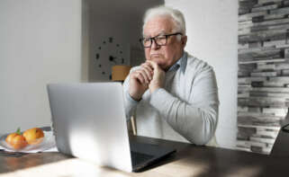 Un adulto mayor sentado en una mesa frente a su ordenador, al cual mira fijamente