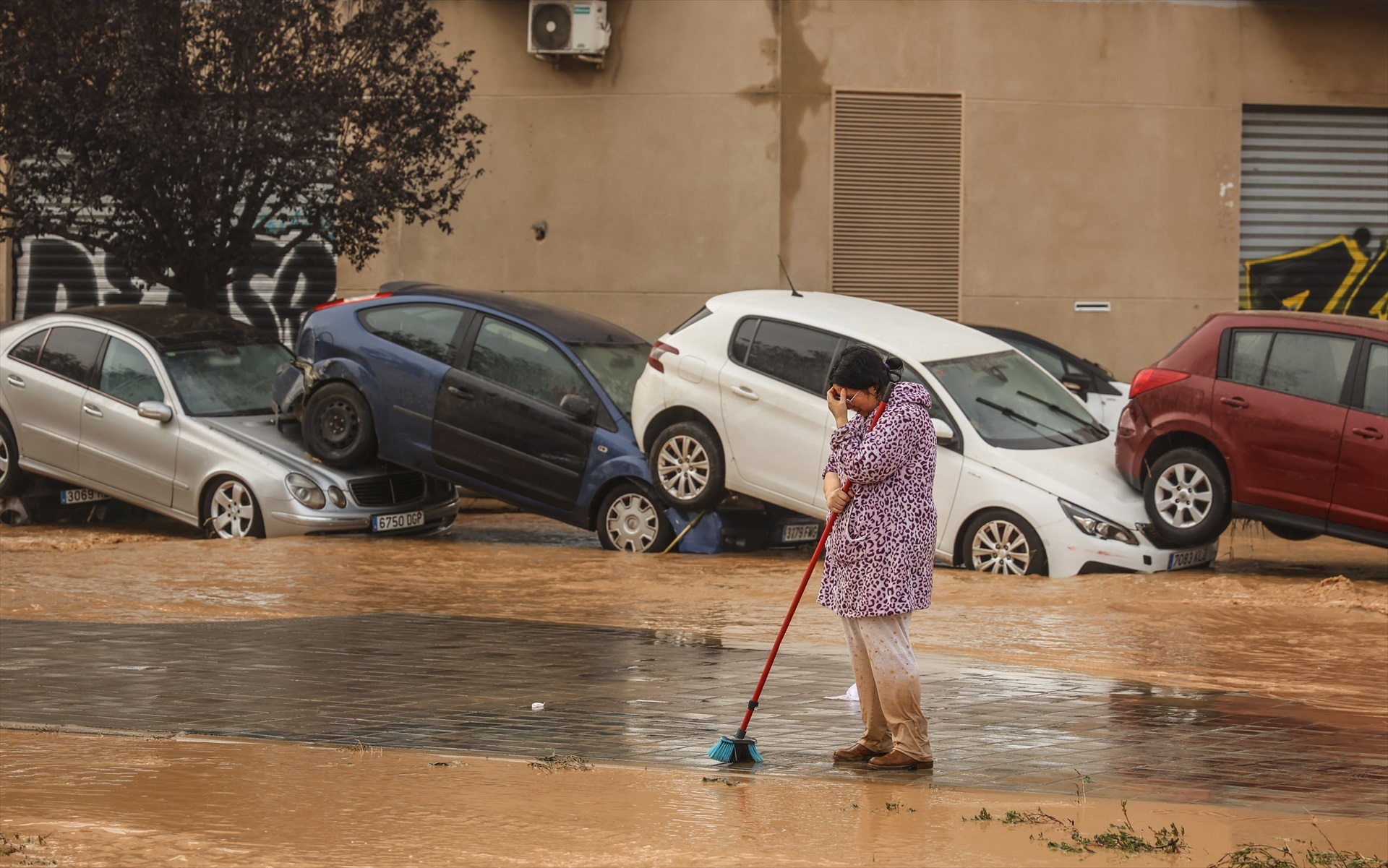 Una mujer realiza labores de limpieza junto a vehículos destrozados tras el paso de la DANA
